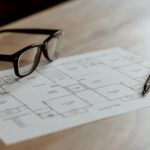 From above of eyeglasses near pen on plan placed on wooden table in apartment in soft daylight