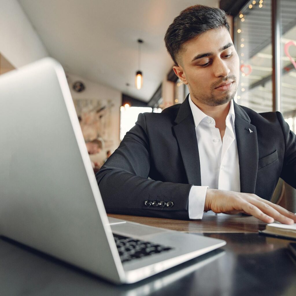 From below of concentrated ethnic male in formal wear sitting in creative workplace and taking notes while using laptop and working on business project