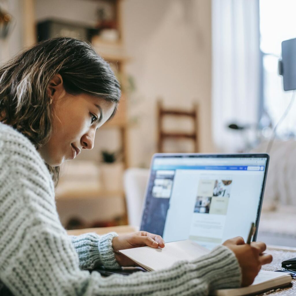 Side view of concentrated young Latin American female freelancer in casual clothes using laptop while taking notes in notebook with pen at table near ring light with phone in room
